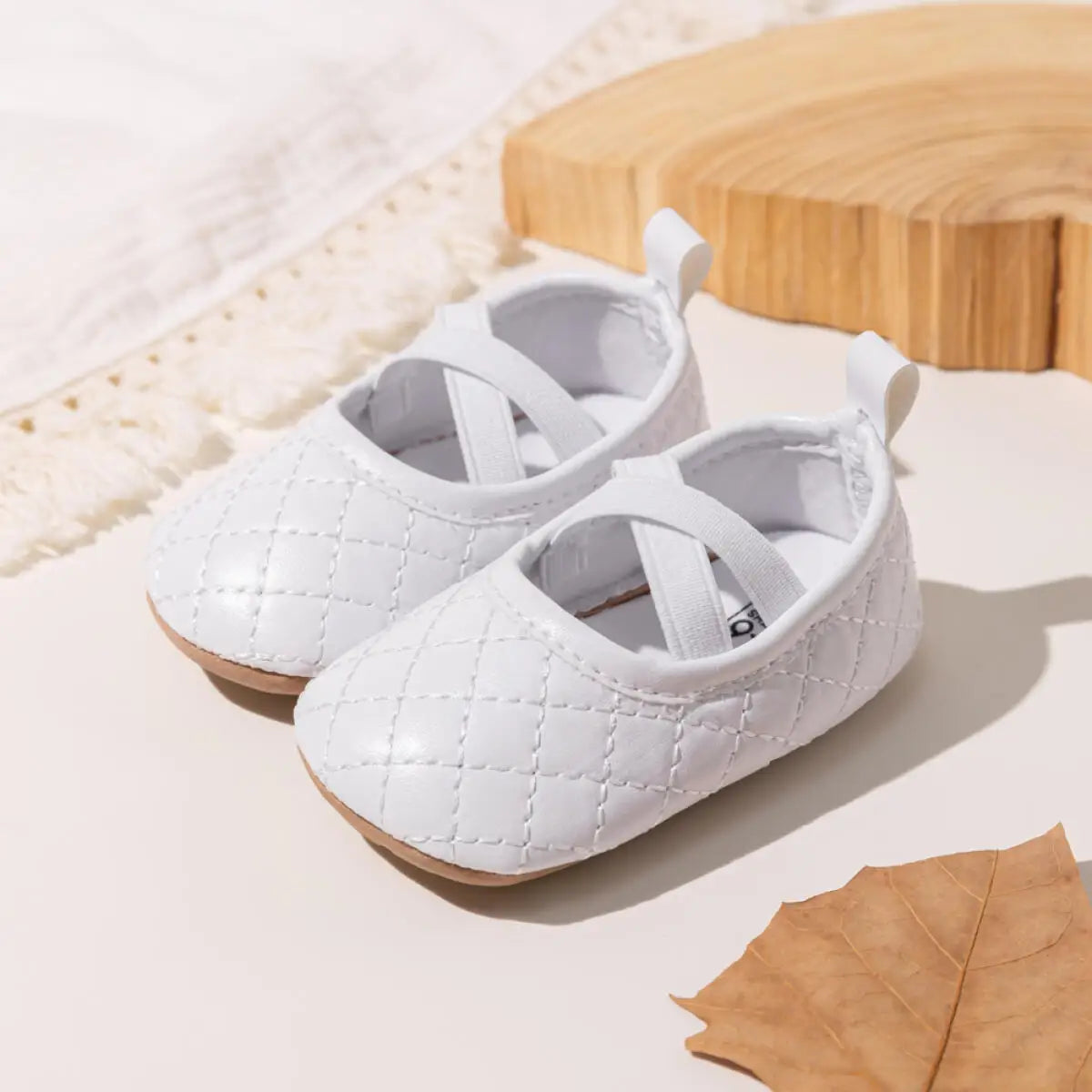 A pair of Kidsely white quilted baby girl ballet flats arranged beautifully with a decorative leaf and wooden blocks.