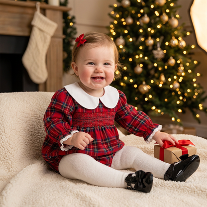 Baby girl wearing red plaid smocked holiday Christmas romper sitting near a holiday tree