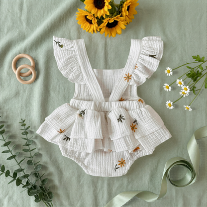 Flat lay showing the detailed ruffle butt backside of an embroidered baby girl romper surrounded by sunflowers, daisy sprigs, and a green ribbon.