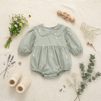 Overhead flat lay view of the sage green daisy embroidered baby bubble romper styled on a cream fabric background with knit booties, a wooden rattle, chamomile flowers, and eucalyptus stems.