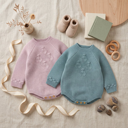 Side-by-side flat lay of light pink and teal baby girl seamless knit sweater rompers with soft accessories.
