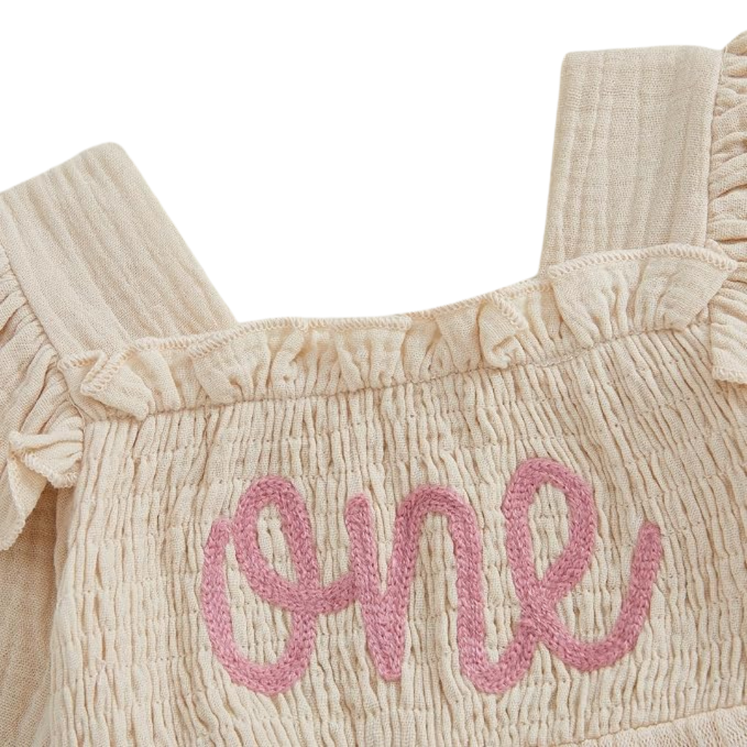 Close up of smocked bodice on cream baby birthday dress with pink embroidered ONE lettering and ruffled shoulder straps