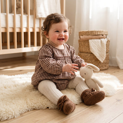 Baby girl sitting in a nursery wearing brown long sleeve smocked gingham top and bloomer outfit set playing with a plush toy.