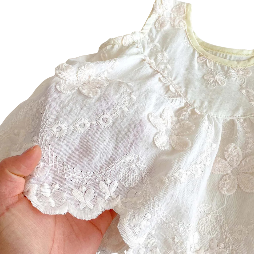 Detailed view of the intricate white lace scalloped hem and embroidered flower patterns on a baby girl’s summer top.