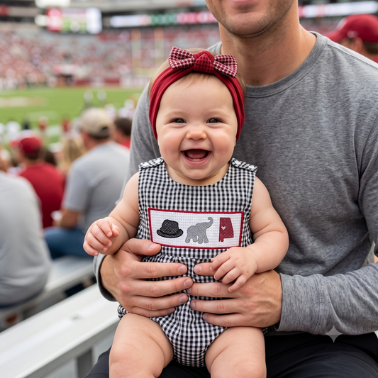 A laughing baby girl wearing a crimson headband with a houndstooth bow sitting with her dad in stadium bleachers, dressed in a black gingham smocked bubble romper featuring an embroidered houndstooth cap, grey elephant, and crimson state outline.
