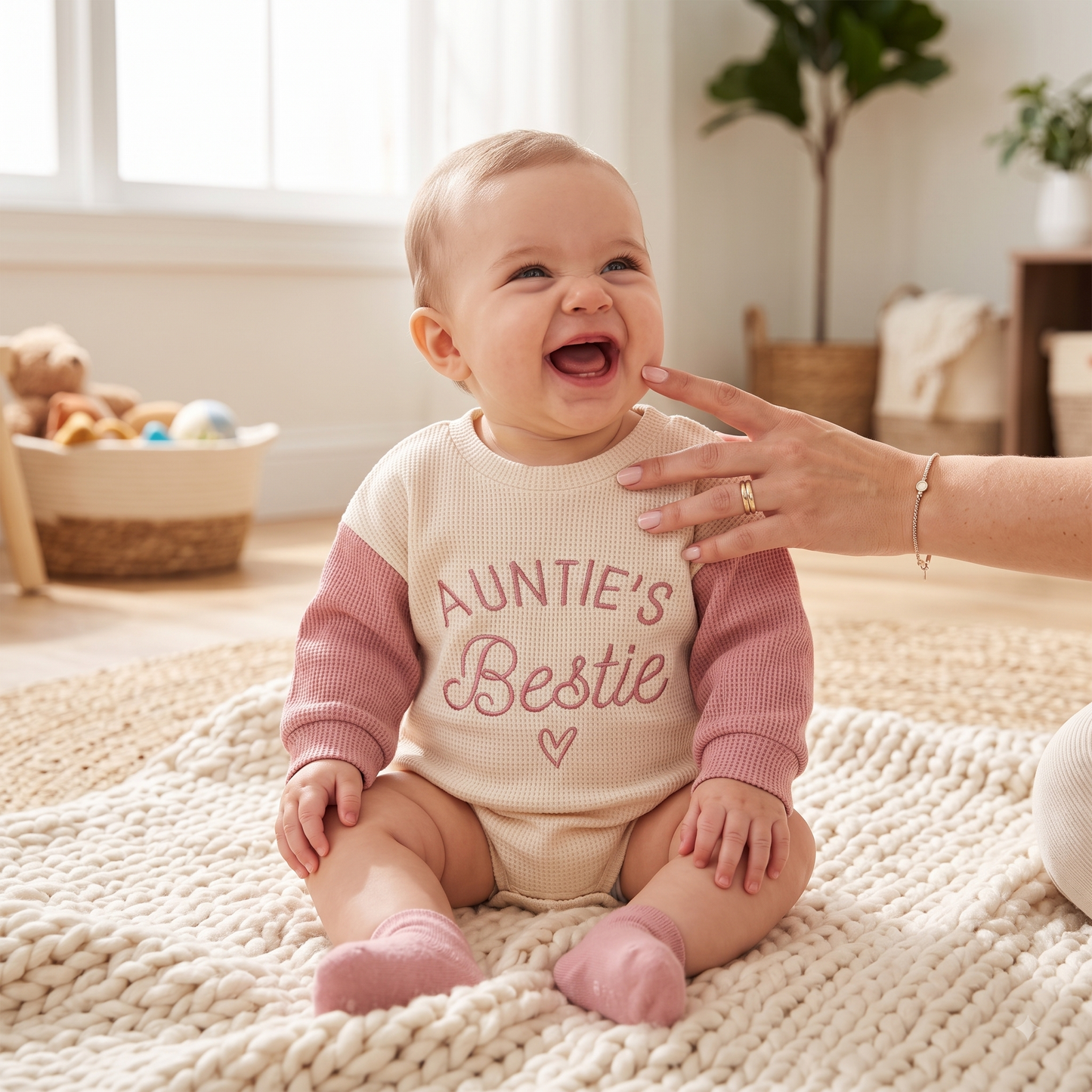 A baby wearing a waffle-knit long-sleeve romper with pink sleeves and a cream body, featuring the text 'AUNTIE'S Bestie' in pink. The baby is sitting on a chunky knit rug in a bright, cozy room, with a hand gently touching the baby's face.