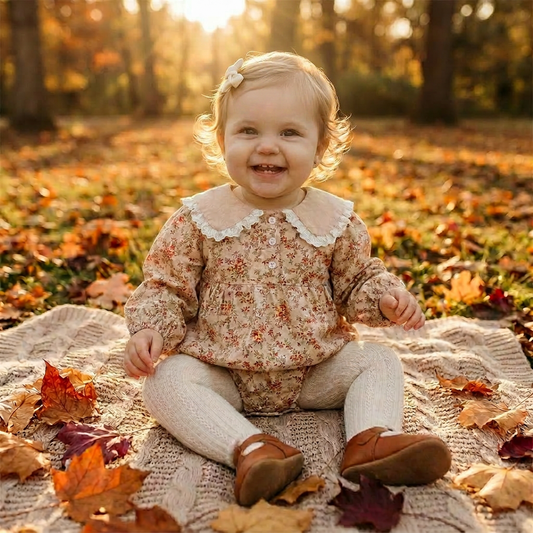 Smiling baby girl sitting outdoors on a blanket surrounded by autumn leaves, wearing the cream vintage floral cottagecore bubble romper.