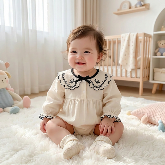 Smiling baby girl wearing an ivory and black long sleeve embroidered bubble romper with matching socks, sitting on a fluffy nursery rug.