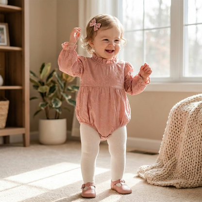 Smiling baby girl standing and modeling a pink floral embroidered bubble romper with long bishop sleeves.