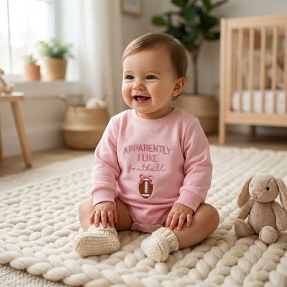 A happy, smiling baby girl sitting on a textured nursery rug wearing the pink long sleeve football bubble romper.