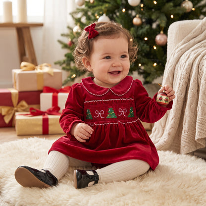 Smiling baby girl wearing a red long sleeve smocked holiday dress sitting next to Christmas presents and a tree.