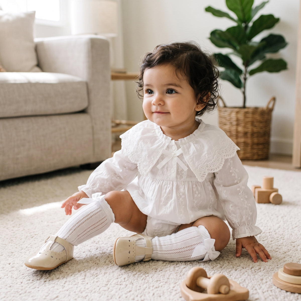 Baby girl sitting on the floor wearing a vintage-style white long-sleeve lace bubble romper with knee-high socks.