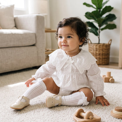 Baby girl sitting on the floor wearing a vintage-style white long-sleeve lace bubble romper with knee-high socks.
