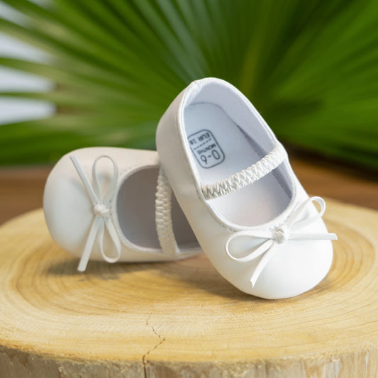 Kidsely white baby girl ballet flat crib shoes with bows on a wooden surface with a green leafy background