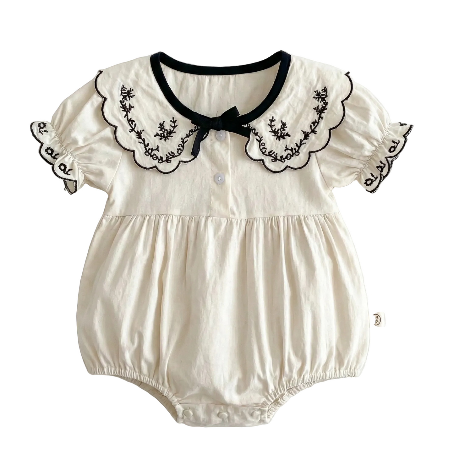 Flat lay of an ivory colored 100% cotton baby girl bubble romper featuring a black embroidered scalloped lapel collar, short puff sleeves, and a black bow detail on a clean white background.