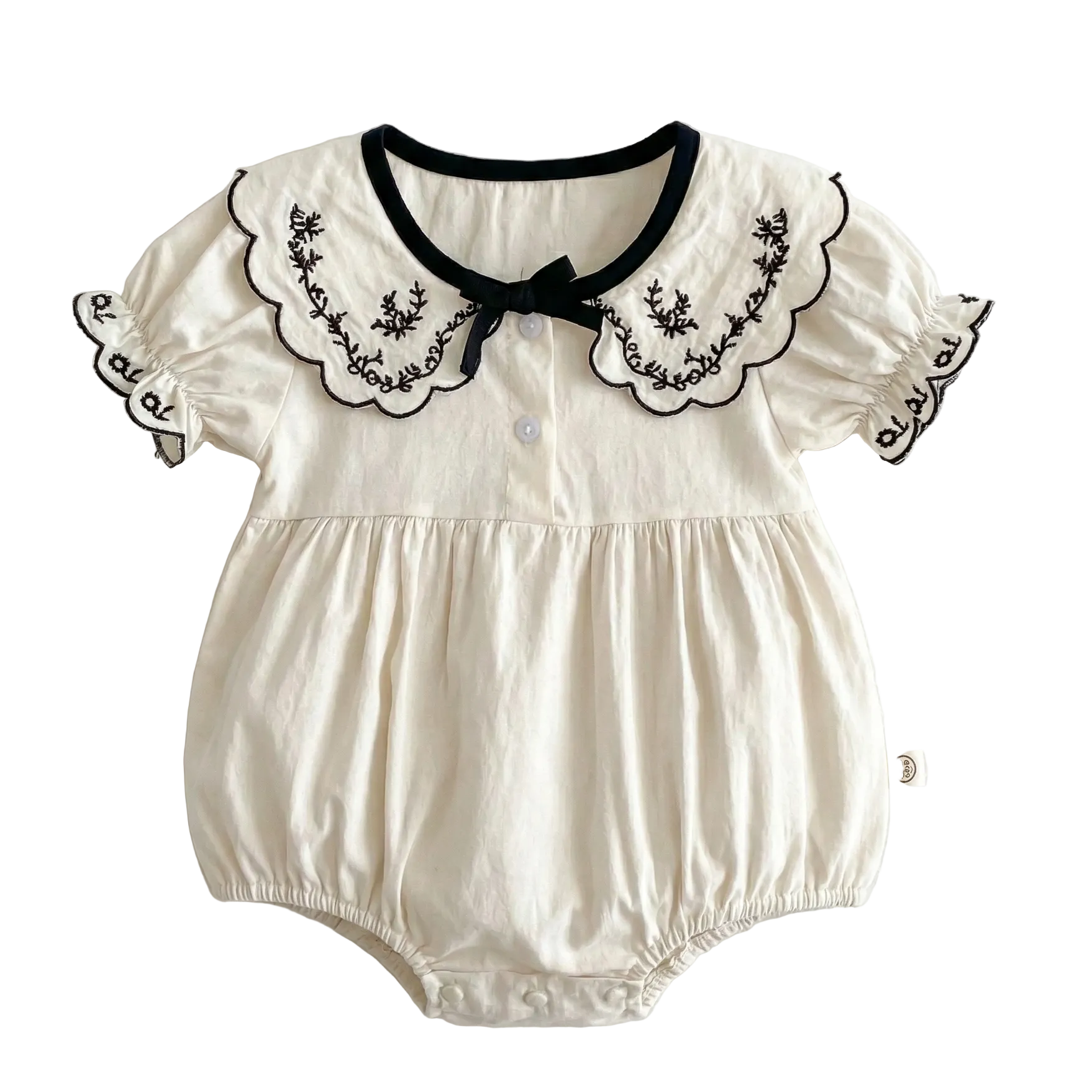 Flat lay of an ivory colored 100% cotton baby girl bubble romper featuring a black embroidered scalloped lapel collar, short puff sleeves, and a black bow detail on a clean white background.