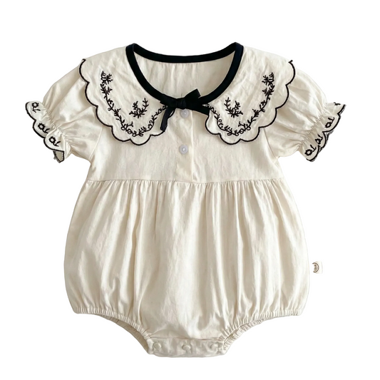 Flat lay of an ivory colored 100% cotton baby girl bubble romper featuring a black embroidered scalloped lapel collar, short puff sleeves, and a black bow detail on a clean white background.