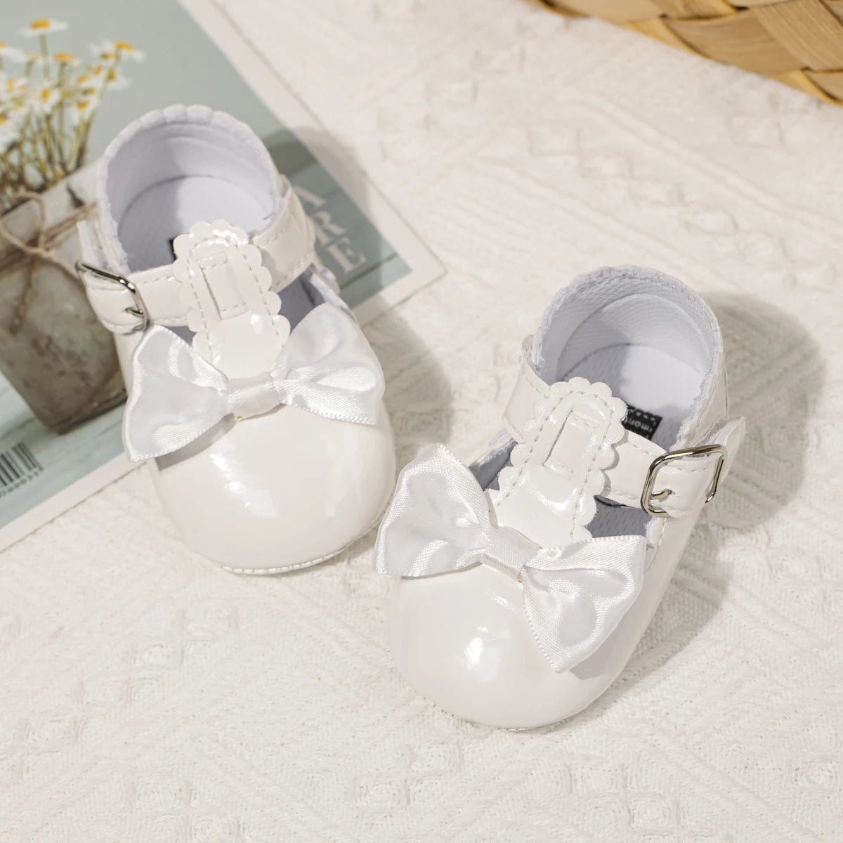 White patent leather baby dress shoes sitting next to a soft floral background, perfect for holiday or baptism outfits.