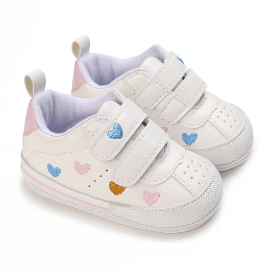 Pair of white baby girl soft sole sneakers featuring multi-colored embroidered hearts, double adjustable straps, and a light pink heel accent.