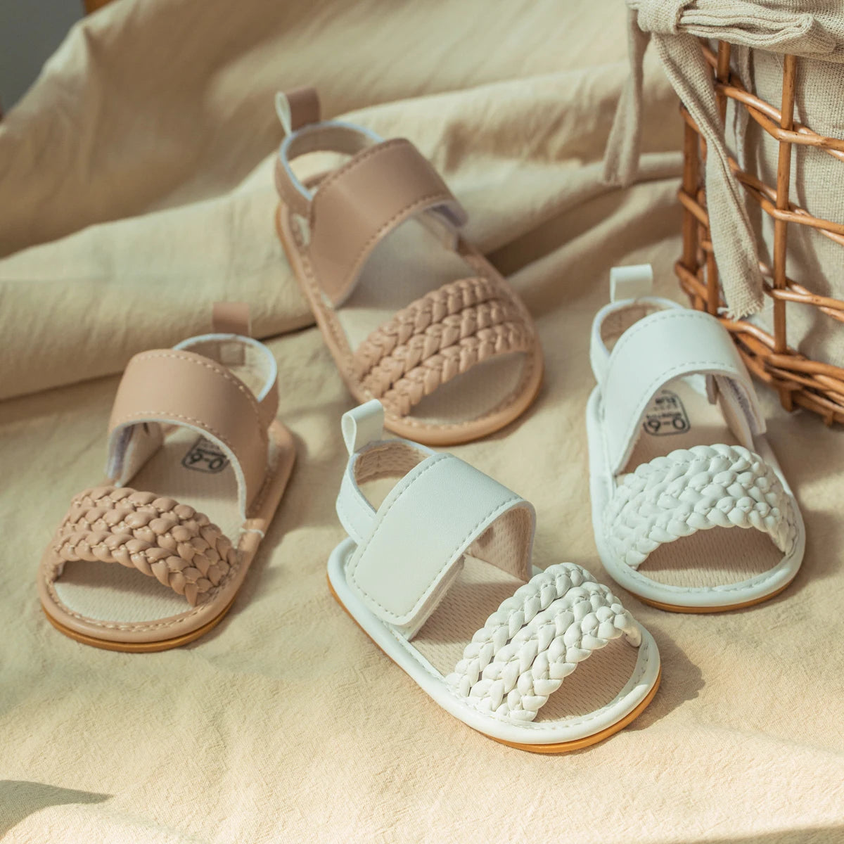 Pairs of white and beige baby girl woven faux leather sandals resting on a blanket next to a woven basket.