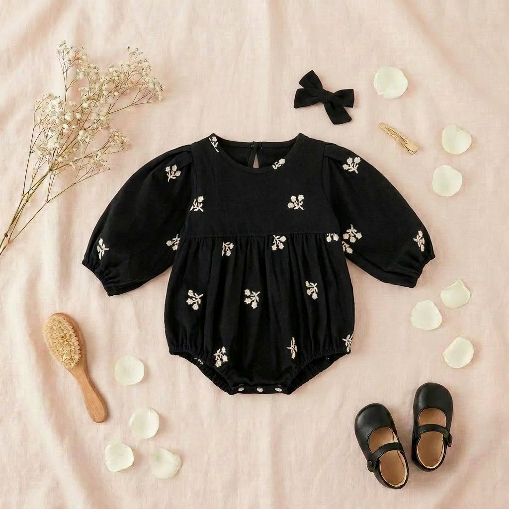 Flat lay of a baby girls black floral embroidered puff sleeve romper on a soft pink background, styled with a black hair bow and baby shoes.