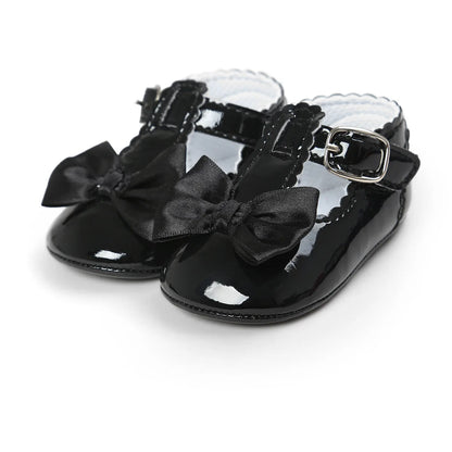 Black patent leather T-strap Mary Jane dress shoes for baby girls featuring a satin bowknot and an adjustable buckle strap on a clean white background.