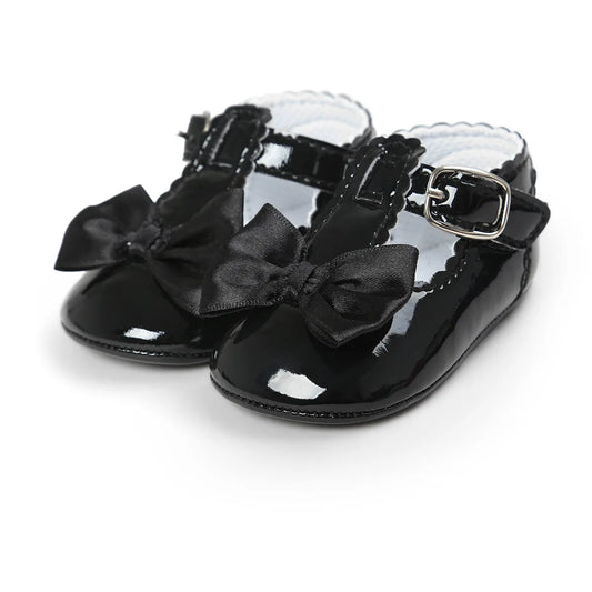 Black patent leather T-strap Mary Jane dress shoes for baby girls featuring a satin bowknot and an adjustable buckle strap on a clean white background.