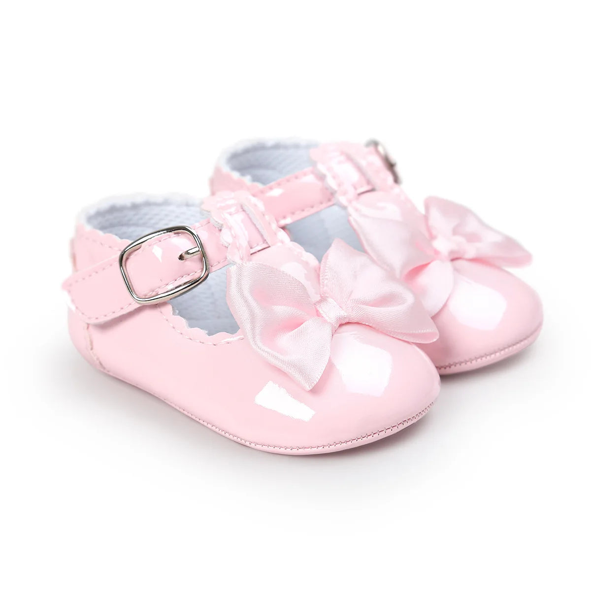 Soft pastel pink patent leather baby girl T-strap Mary Janes with a matching pink satin decorative bow and silver buckle.