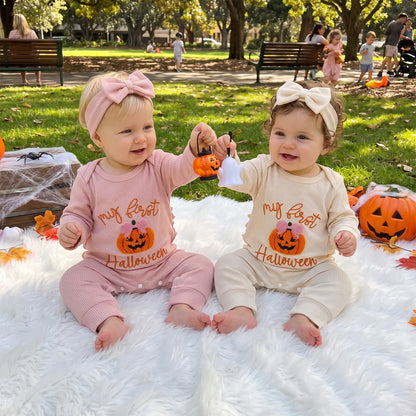 Two smiling baby girls sitting outdoors on a white blanket wearing matching pink and cream "My first Halloween" rompers with headbands.
