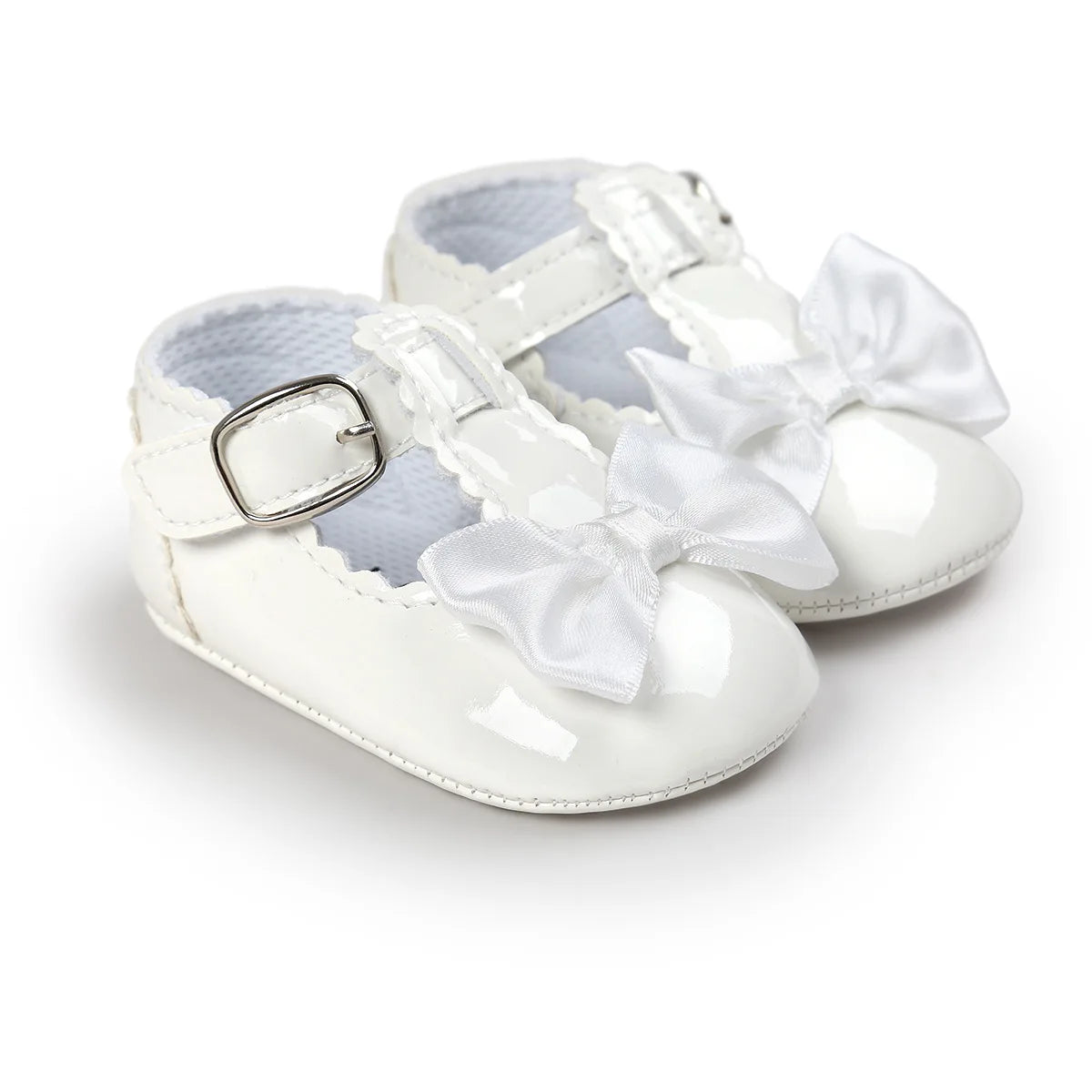 A pair of white patent leather baby girl T-strap Mary Jane shoes with a white satin bow and metallic silver buckle buckle closure.