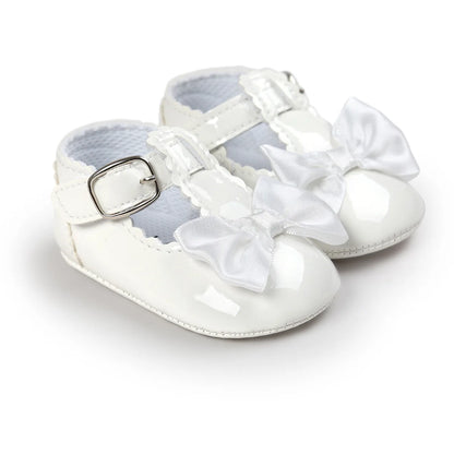 A pair of white patent leather baby girl T-strap Mary Jane shoes with a white satin bow and metallic silver buckle buckle closure.