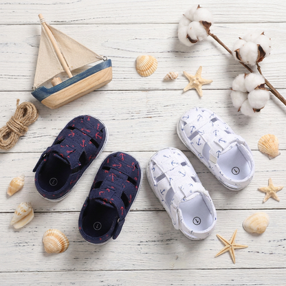 A nautical themed flat lay featuring navy and white baby anchor print canvas sandals styled with a toy wooden boat and seashells.