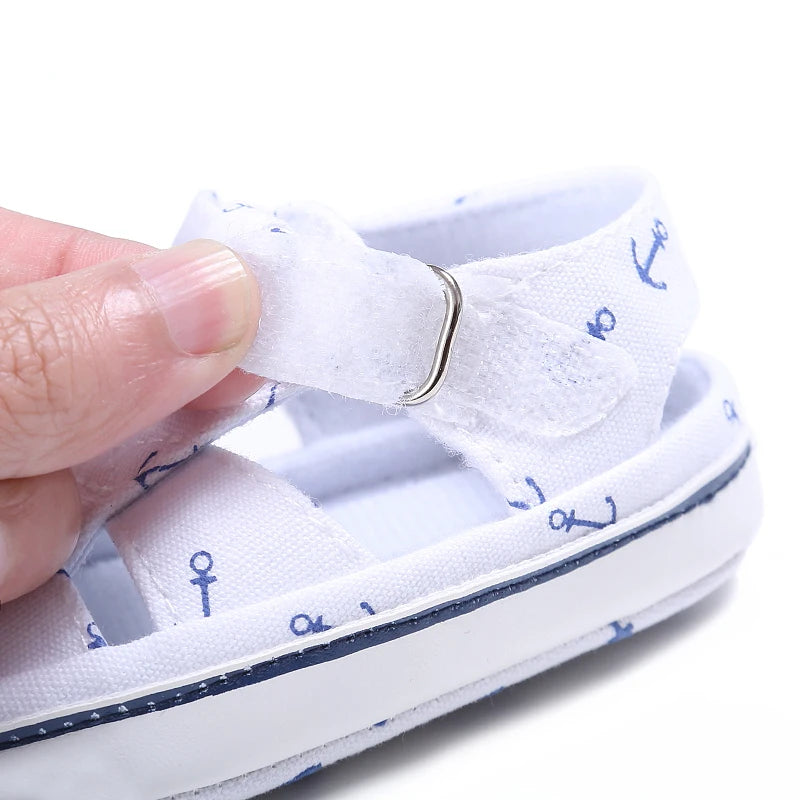 Close-up of a hand opening the adjustable hook-and-loop velcro strap on a white nautical baby sandal.