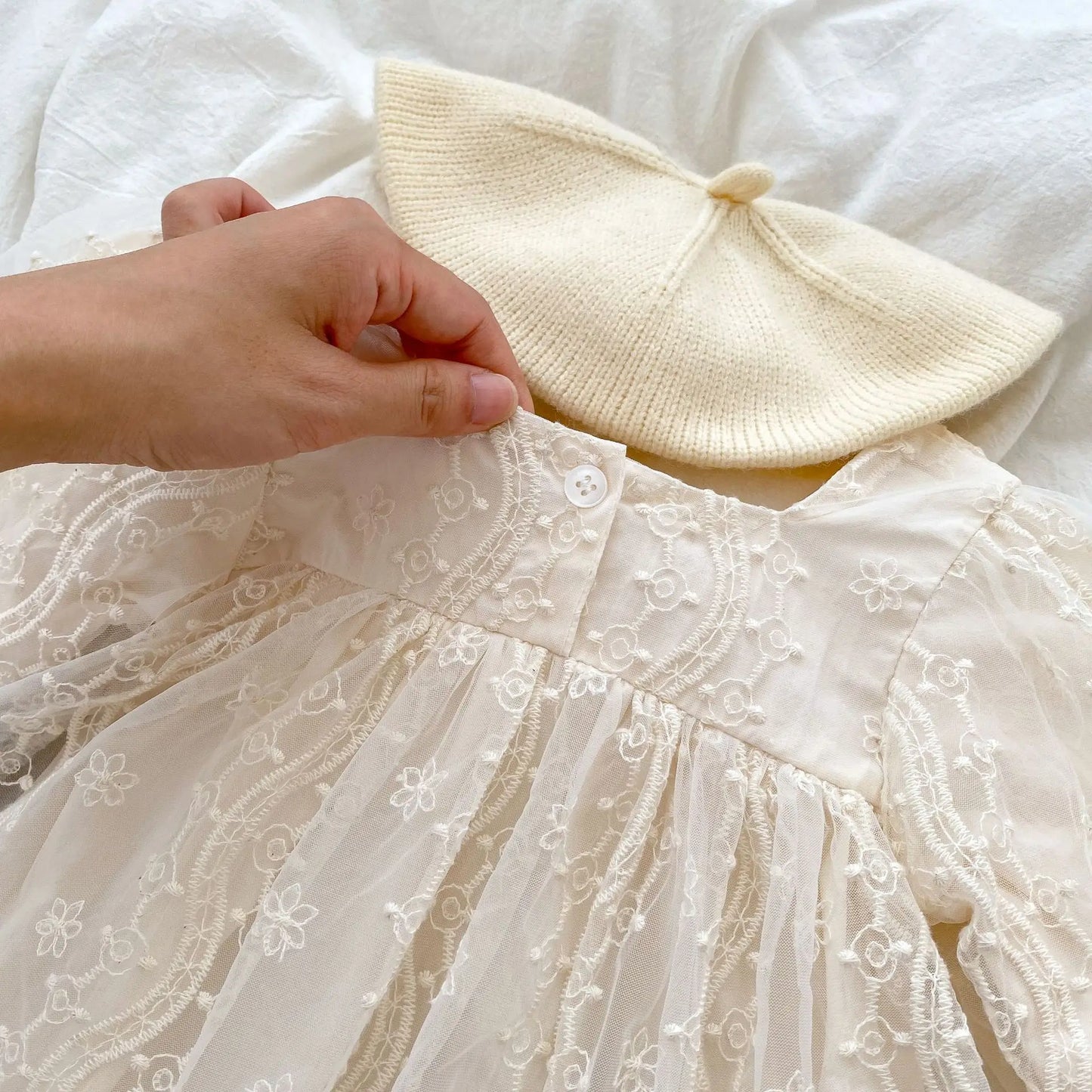 Close-up of the back neckline of a baby girl lace romper showing the top button closure, next to a cream knit beret.
