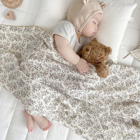 Sleeping baby comfortably tucked under a lightweight taupe botanical muslin swaddle blanket next to a teddy bear.