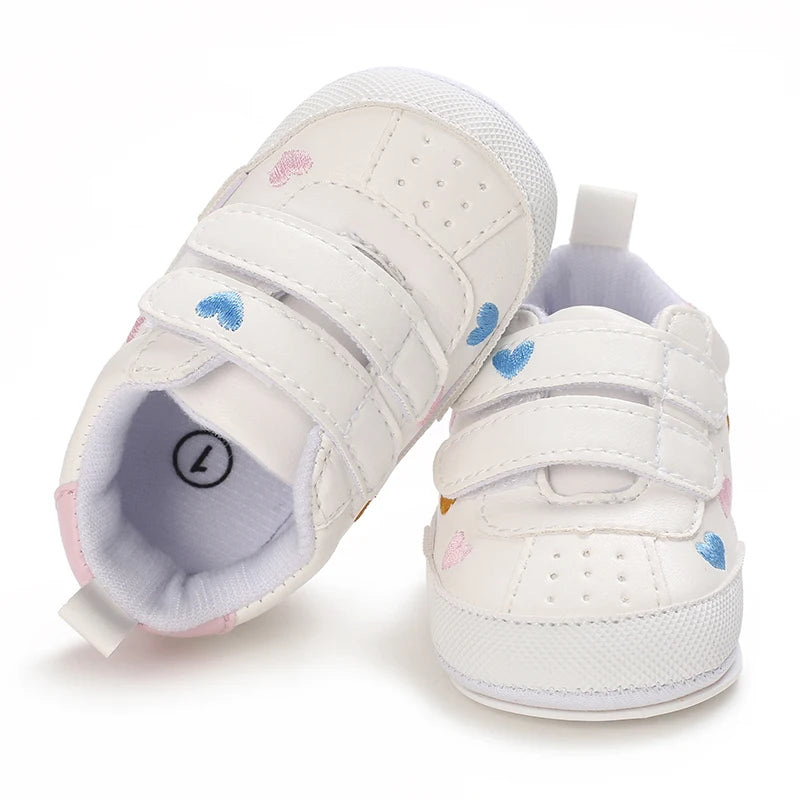 Top-down view of white infant sneakers with heart prints, showing the open double hook-and-loop straps and soft interior lining.