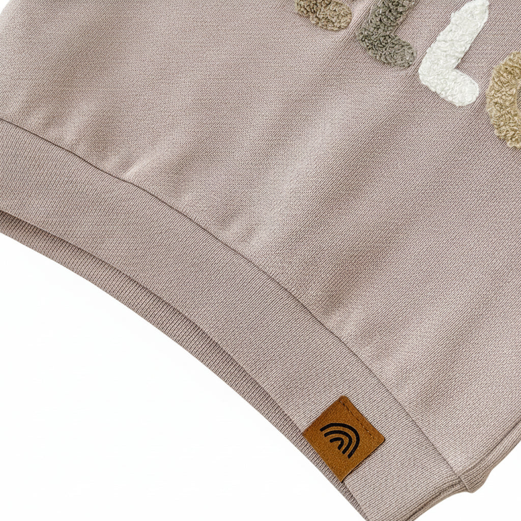 Close-up of the ribbed bottom hem on the beige toddler pullover top featuring a small brown tag with a rainbow logo.
