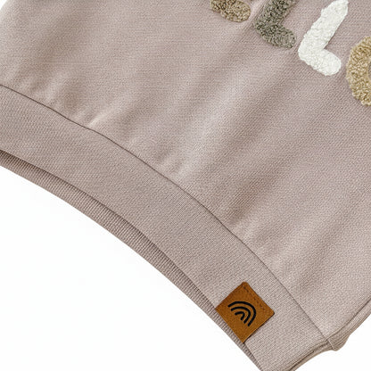 Close-up of the ribbed bottom hem on the beige toddler pullover top featuring a small brown tag with a rainbow logo.