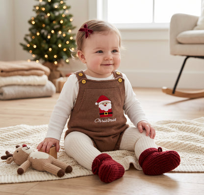 A smiling baby sitting on a rug wearing the brown Santa Claus knit romper layered over a white long-sleeve shirt and red booties, with a toy reindeer and a small Christmas tree in the background.