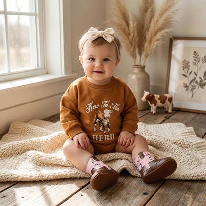 Smiling baby wearing the rust New To The Herd western long sleeve bubble romper sitting on a blanket.