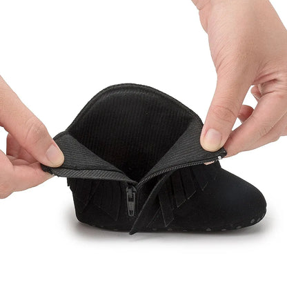 Hands bending a black infant fringe suede moccasin bootie to demonstrate the ultra-flexible first walker construction.