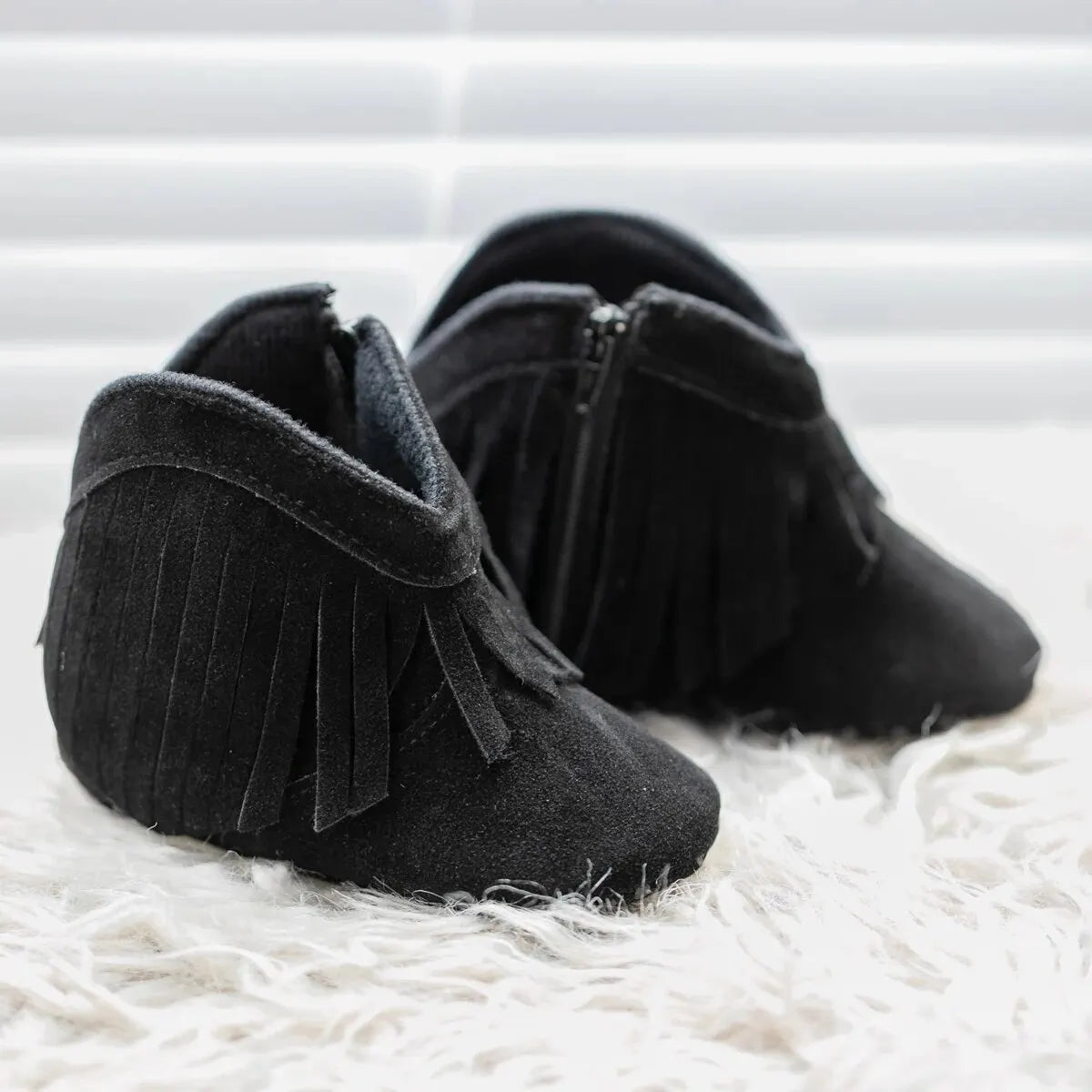 Pair of black baby western fringe moccasin booties, front view on a soft white textured rug.