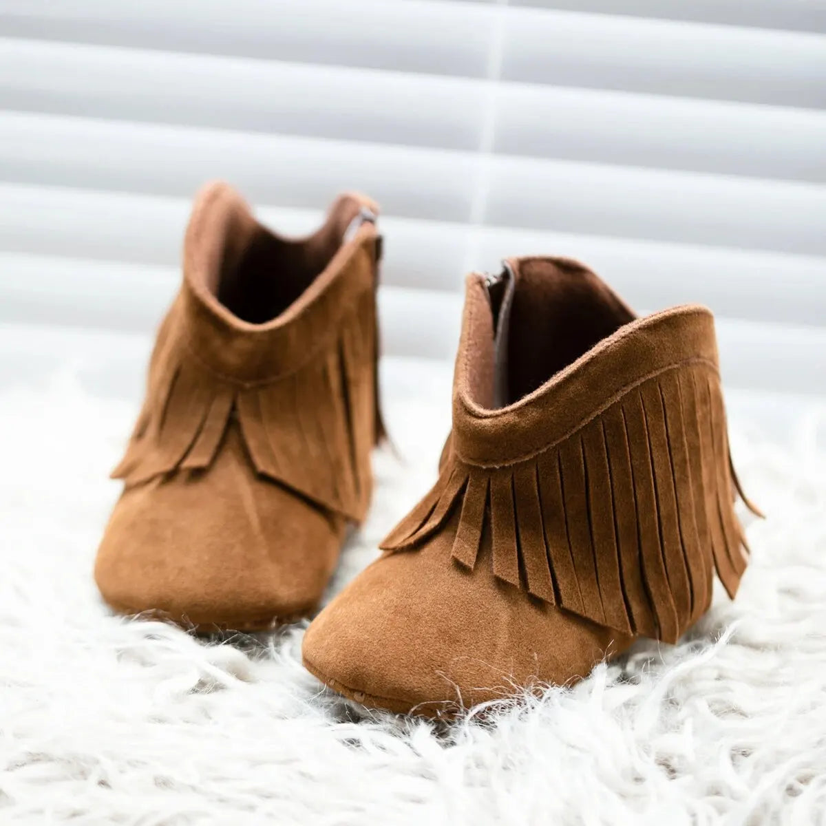 Angle view of brown infant fringe suede booties resting on a white surface next to a window.