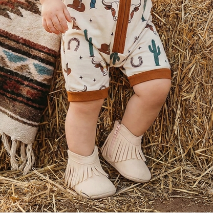 Close up of an infant wearing cream baby western fringe moccasin booties and a western-printed romper with longhorns and cacti, standing by a hay bale.