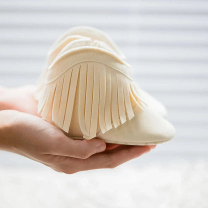 A person holding a cream-colored soft sole baby fringe moccasin bootie to display its premium tassel details
