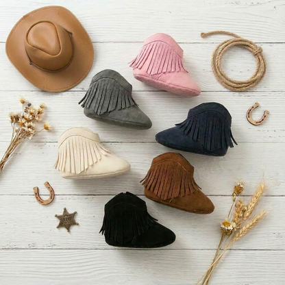 Overhead flat lay of Kidsely baby western fringe moccasin booties in pink, cream, gray, navy, brown, and black on a white wooden surface with a miniature cowboy hat, rope lasso, and sheriff star badge.