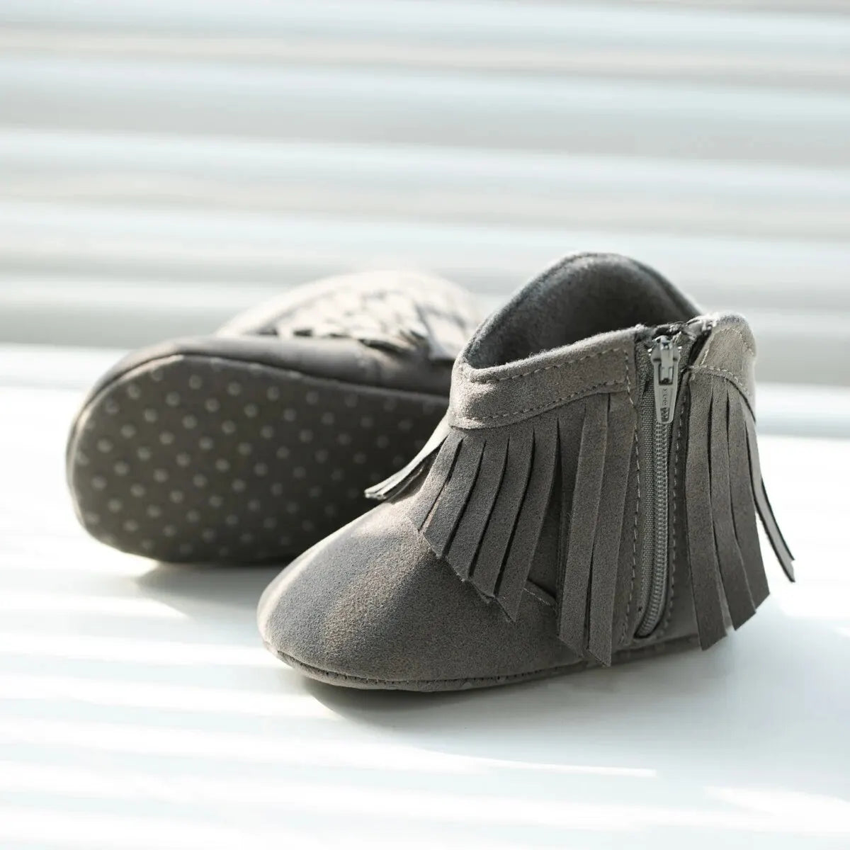 Side profile view of gray baby western fringe moccasin booties showing the side zipper next to a bright window.