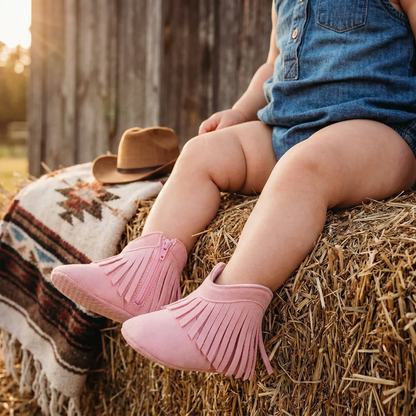 An infant wearing pink baby western fringe moccasin booties and a denim romper, sitting on a hay bale next to a mini cowboy hat and southwestern blanket.
