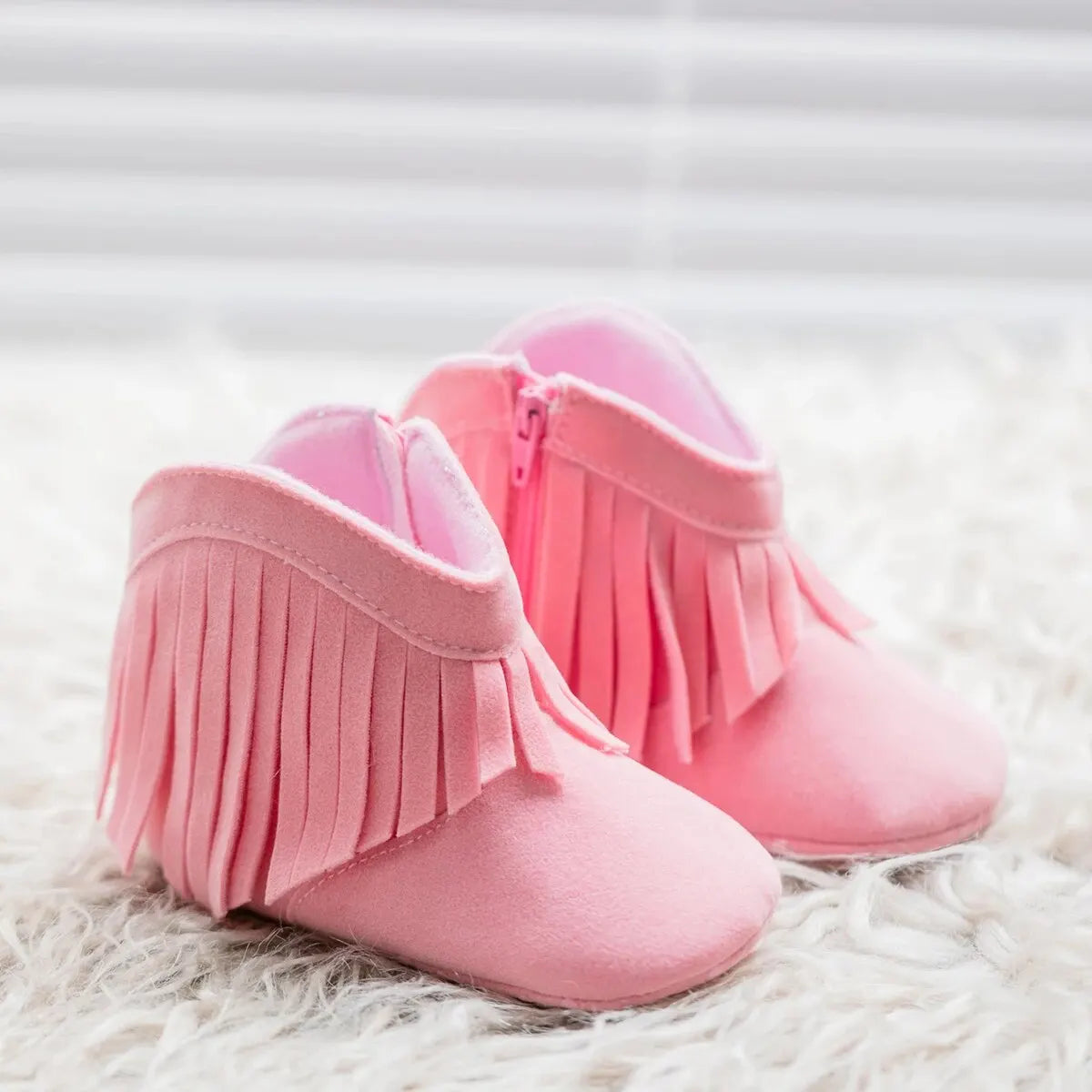 Side profile view of pink infant fringe suede booties showing the side zipper closure on a soft white fur surface.