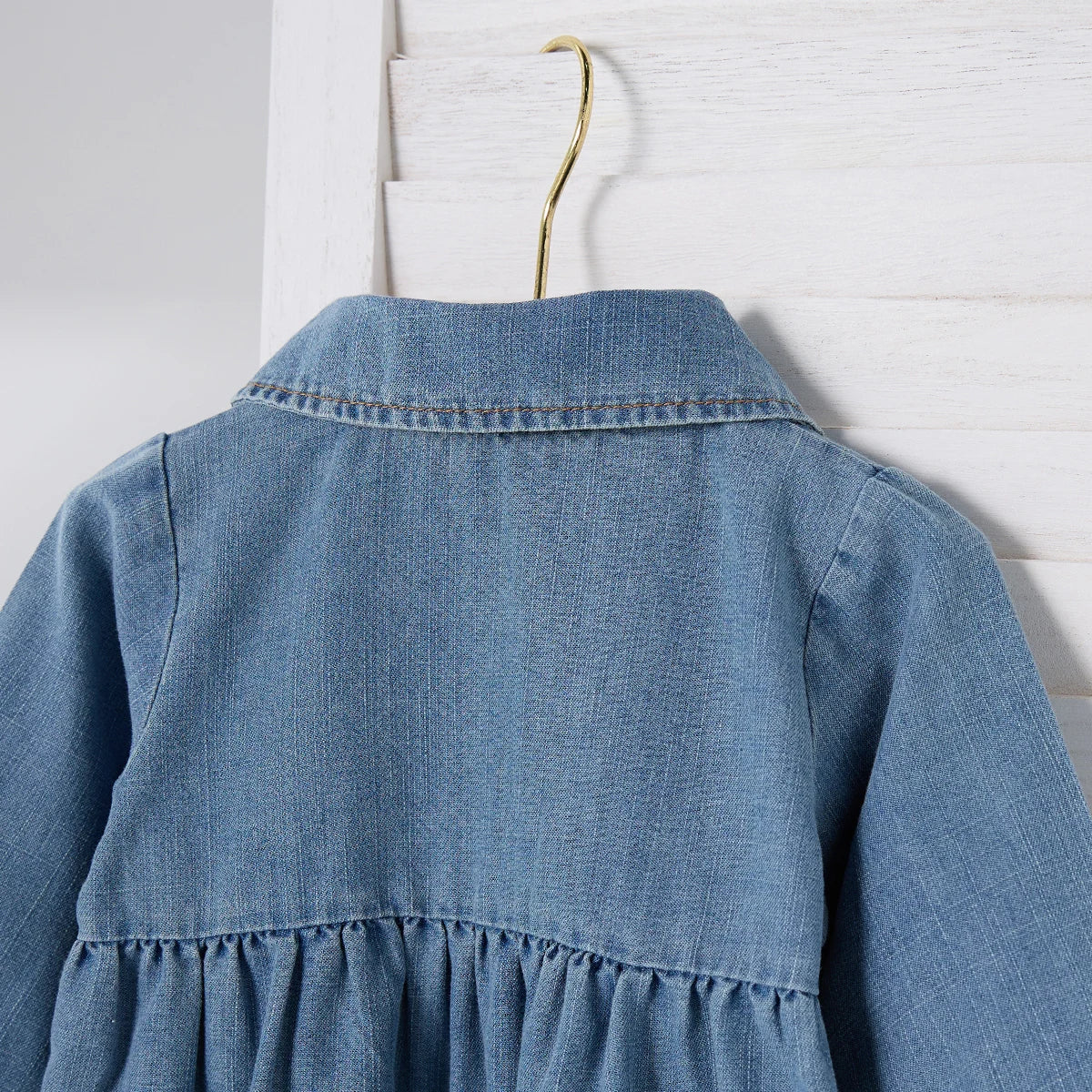 Close-up of the back shoulder and gathered waist detail on a blue denim playsuit hanging on a hanger.